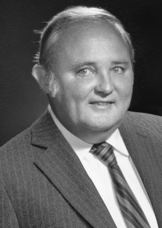 Black and white portrait of an older man wearing a pinstripe suit, striped tie, and white shirt, smiling slightly and facing the camera against a dark studio background.
